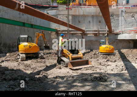 Un lavoratore libera le catene dalla gru che si è abbassato di un gatto caricatore cingolato compatto sulla sua area di lavoro. Sullo sfondo due JCB escavatori compatti. Foto Stock