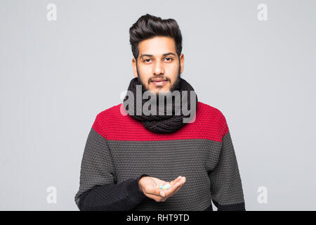 Ritratto di un uomo indiano vestito con un maglione e sciarpa che mostra una pillola capsula in piedi su sfondo grigio Foto Stock