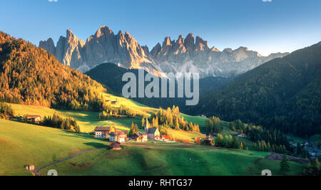 Paesaggio con villaggio con case, la chiesa, il verde dei prati, alberi, rocce, cielo blu. Santa Maddalena in Valle di montagna al tramonto in autunno nelle Dolomiti Foto Stock