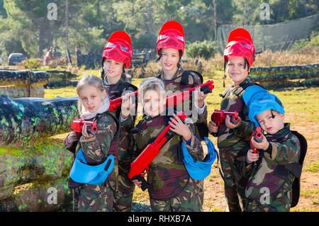 Gentile lieto allegro sorriso positivo gruppo di bambini paintball giocatori in camuffamento in posa con pistole di paintball sul campo da gioco all'aperto Foto Stock