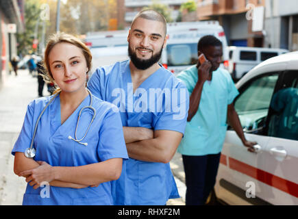 Due positive cortese ed efficiente paramedicals fiducioso in uniforme in posa in prossimità di ambulanza Foto Stock