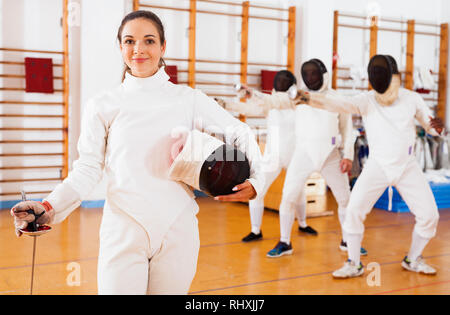 Sorridendo allegro sportivo positivo giovane donna in uniforme in piedi in allenamento di scherma Foto Stock