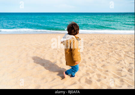 Close up lonely baby boy camminare da solo sulla spiaggia di sabbia durante la giornata di vento andando via Foto Stock