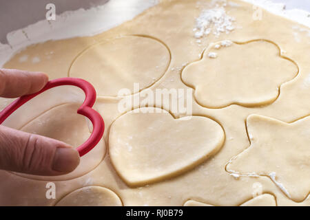 Portata di Mano premendo una a forma di cuore ad cookie cutter in un foglio di zucchero pasta biscotto. Tagliare forme cookie includono: cuore, fiore, stelle e cerchio. Cl Foto Stock