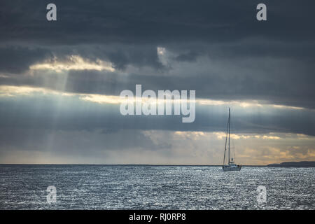 Rainy cielo nuvoloso al di sopra del lone yacht a vela in mare. L'Irlanda del Nord. Sun luci, pioggia fluisce oltre l'acqua di mare. Barca solitaria tra l'oceano. Schiacciante del paesaggio marino in grigio blu tinte. Foto Stock