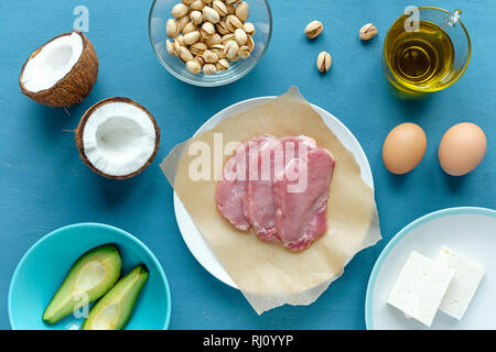 Layout di overhead di ingredienti per la cottura su sfondo blu. Fette di carne cruda per bistecche sulla carta, cocco fresco, avocado, pistacchi, formaggio, uova Foto Stock