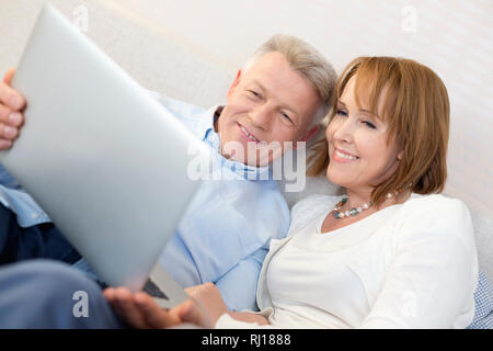 Sorridente Coppia matura utilizzando computer portatile in camera da letto a casa Foto Stock