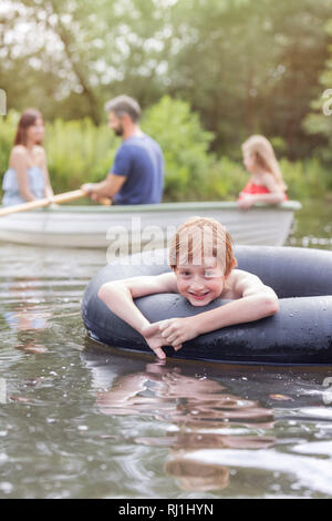 Ritratto di ragazzo sorridente flottante con anello gonfiabile nel lago contro la famiglia Foto Stock