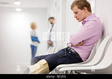 Uomo che guarda il rinforzo sul ginocchio mentre è seduto in ospedale in sala d'attesa Foto Stock