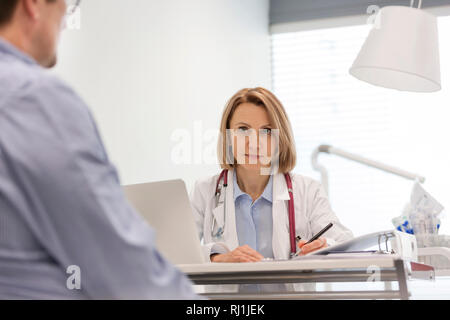 Medico discute con matura paziente durante la scrittura di prescrizione alla scrivania in ospedale Foto Stock