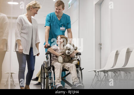 La madre a piedi con un infermiere spingendo il ragazzo malato su una sedia a rotelle in ospedale corridoio Foto Stock