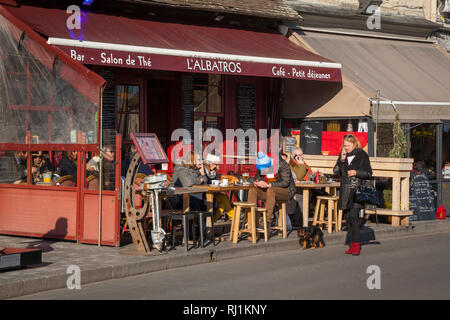 Una signora sul suo telefono passeggiate il suo cane passato un bar su St. Catherine's Quay in Honfleur, Normandia, Francia. Foto Stock