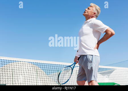 Uomo maturo smorfie con mal di schiena tenendo racchetta da tennis contro il cielo blu e chiaro sulla giornata di sole Foto Stock