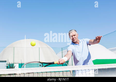 Fiducioso uomo maturo colpendo palla da tennis con la racchetta in tribunale contro il cielo blu chiaro Foto Stock