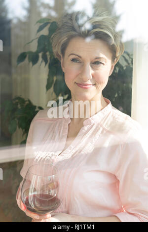 Sorridente donna matura azienda wineglass visto attraverso la finestra Foto Stock