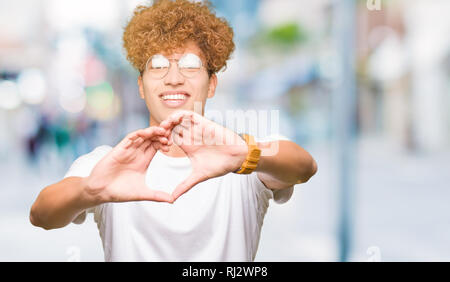 Giovane uomo bello con capelli Afro indossa occhiali sorridente in amore che mostra il simbolo del cuore e la forma con le mani. Concetto romantico. Foto Stock