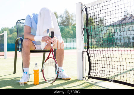 Lunghezza completa di stanco uomo maturo con la testa coperta seduti su una sedia da net al campo da tennis sulla giornata di sole Foto Stock