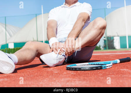 Sezione bassa di uomo maturo stretching gamba mentre è seduto sul rosso campo da tennis durante l'estate Foto Stock