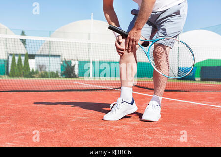 Sezione bassa di uomo maturo holding racchetta da tennis mentre che soffrono di dolore al ginocchio su rosso campo da tennis durante l'estate Foto Stock