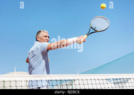Basso angolo di vista fiducioso uomo maturo colpendo palla da tennis con la racchetta in tribunale contro il cielo blu chiaro Foto Stock