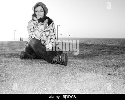 Giovane ragazza in abbigliamento casual e urban hat, felpa con cappuccio e jeans, seduto sul prato in un parco, oltre la visualizzazione del mare, guardando a parte nella contemplazione. Foto Stock