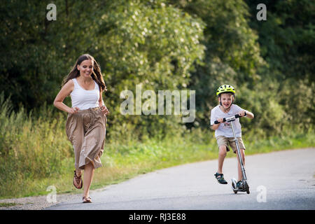 Un piccolo ragazzo scooter di equitazione e madre in esecuzione su una strada nel parco in un giorno d'estate. Foto Stock