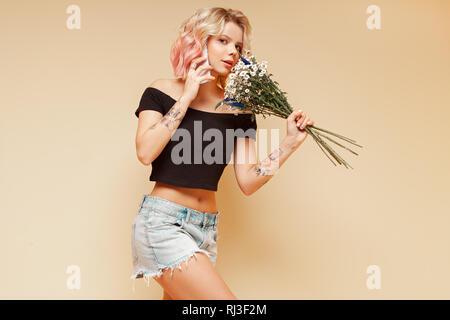 Giovane donna tanga con i colori dei capelli ricci e tatuaggio sorridente e parlare al telefono tenendo i fiori. Foto Stock