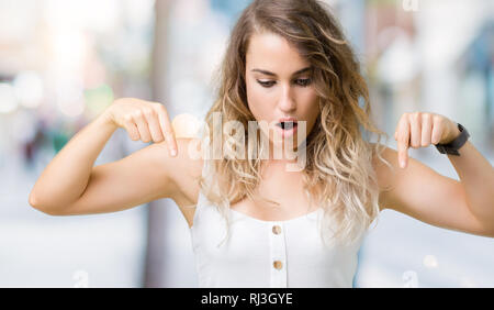 Giovane e bella donna bionda su sfondo isolato con la punta rivolta verso il basso con le dita che mostra annuncio, sorpreso viso e bocca aperta Foto Stock