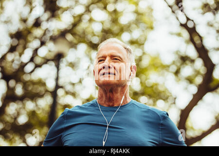 Bassa angolazione di un anziano uomo in piedi sotto un albero per ascoltare musica durante l'allenamento. Close up di un senior persona fitness facendo allenamento all'aperto. Foto Stock