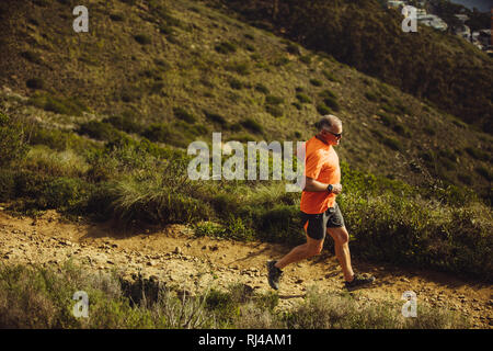 Athletic uomo trail running discesa da una collina in una giornata di sole. Elevato angolo di visione di un uomo anziano in abiti di fitness in esecuzione su una ruvida terreno collinare. Foto Stock