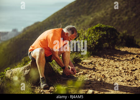 Senior athletic uomo seduto su una roccia accanto a un percorso di prova e legatura laccio. Uomo in usura fitness serrando il suo laccio durante la corsa su sentiero su un Foto Stock