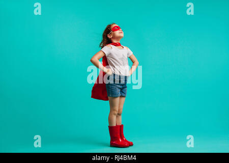 Ragazza indossando gumboots rosso, capo e la maschera per gli occhi in piedi con le mani sui fianchi in studio. Super ragazza guarda lontano su sfondo blu. Foto Stock