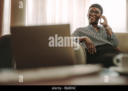 Matura imprenditrice africana in viaggio d'affari seduti sul divano e parlando al telefono in camera d'albergo. Donna in abiti formali seduto in camera di albergo e ma Foto Stock