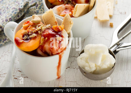 Gustosi piatti freddi appetitosi gelato alla vaniglia con dadi, albicocche, cialde e sciroppo sul tavolo bianco con gli ingredienti per la produzione di dessert. Primo piano. Foto Stock