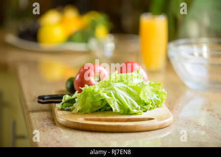 Tagliere di legno sul tavolo con verdure fresche, succo d'arancia sullo sfondo Foto Stock