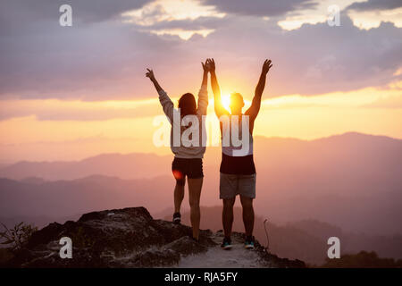 Coppia felice con i bracci sollevati sorge sulla cima della montagna contro il tramonto e divertirsi in vincitore pongono Foto Stock