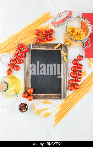 Piccola scheda di gesso con copia spazio con gli ingredienti per la pasta italiana La cucina e i pomodori ciliegia sulla tavola bianca, vista dall'alto Foto Stock