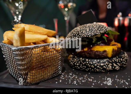 Nero gustosi hamburger e patatine fritte nel cestello servita su nero tagliere. Messa a fuoco selettiva. Copia-bandiera dello spazio bloccato in bun per il testo individuale. Foto Stock