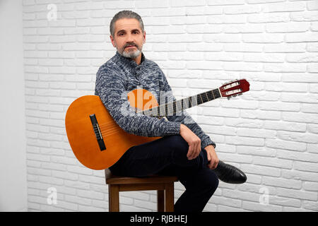 Bello barbuto uomo seduto su una sedia in studio e guardando la fotocamera. Uomo maturo tenendo la chitarra acustica. Fiducioso musicista in posa con lo strumento musicale. Foto Stock