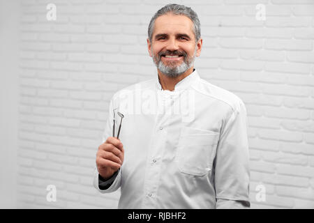 Felice dentista in posa di studio con sfondo bianco. Bello stomatologist indossa in bianco uniforme del laboratorio di restauro di contenimento degli strumenti, guardando la fotocamera e sorridente. Foto Stock