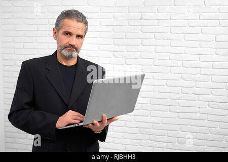 Gravi e fiducioso imprenditore in posa di studio con sfondo bianco. Bel uomo maturo guardando la telecamera, tenendo notebook. Uomo Barbuto che indossa in abito nero. Foto Stock