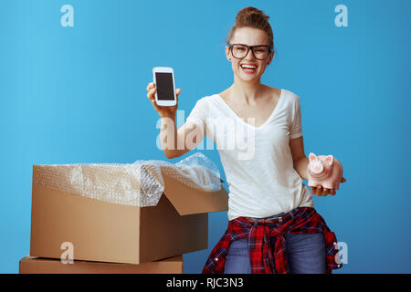 Sorridente giovane donna in t-shirt bianca con piggybank mostra Telefono nella schermata vuota vicino alla scatola di cartone isolato sul blu. Chiamare professional movers e ott Foto Stock