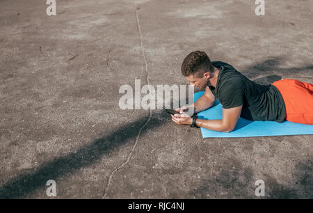 Giovane ragazzo su rug, nelle mani di smartphone. Guardare video motiva per attività sportive. Visualizzare le calorie in applicazione tramite Internet. Concetto di Foto Stock