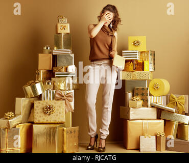 Sottolineato eleganti donna in oro pantaloni beige e marrone camicia con notebook dorata che ricopre la faccia con le mani tra 2 pile di golden doni nella parte anteriore di un Foto Stock