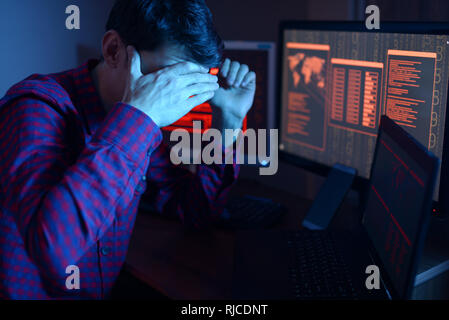 Un anonimo hacker maschio si copre il viso con la mano mentre commette un crimine di hack il sistema sullo sfondo delle schermate nel buio. Il concetto o Foto Stock