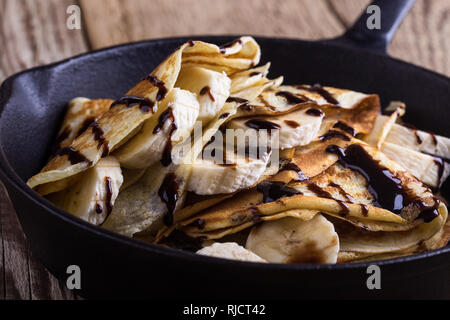 Crepes fatte in casa con banane e topping al cioccolato per colazione in padella in ghisa su tavola in legno rustico, vicino il fuoco selettivo Foto Stock