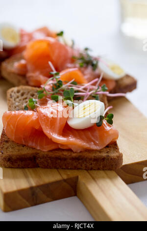 Toast con salmone affumicato e uovo di quaglia e germogli di rafano servito su rustiche di legno con bicchieri di vino bianco. Deliziosi aperitivi e snack o pa Foto Stock