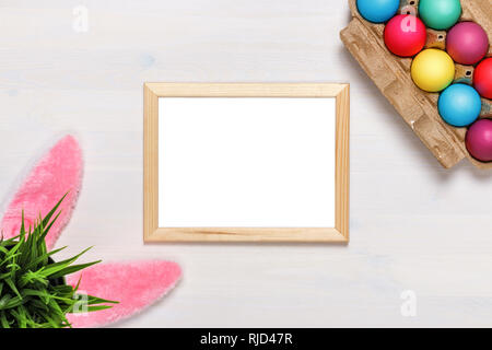 Un telaio, orecchie di coniglietto, un vaso di fiori artificiali con erba verde e colorati di uova in una scatola di cartone, vernici e pennelli. Concetto di pasqua. Spazio di copia Foto Stock