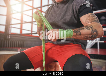 Preparazione di combattimento. Atletica muscolare uomo mani di incarto con verde avvolge boxe boxe in palestra, close-up di forti mani e pugno pronto per la lotta Foto Stock