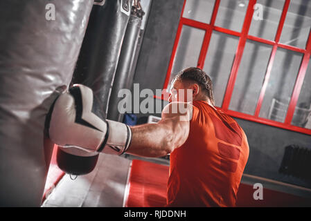 Close-up di un forte colpo al punzone in borsa. Fiducioso sportman muscolare in guanti bianchi allenamento duro sul punzone pesante sacco nero in palestra di inscatolamento Foto Stock